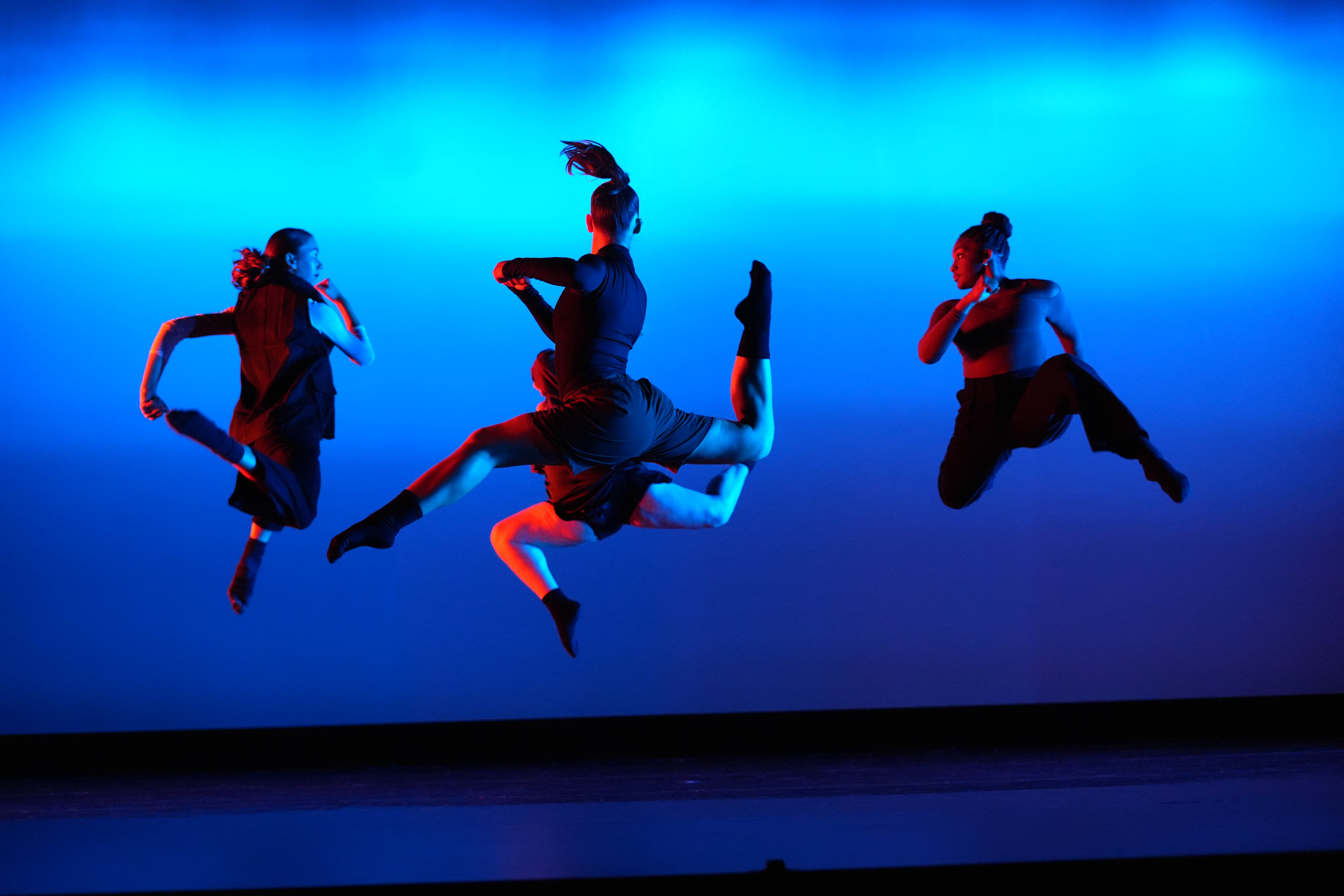 Four dancers jumping in mid-air, circling each other, against a blue-lit backdrop