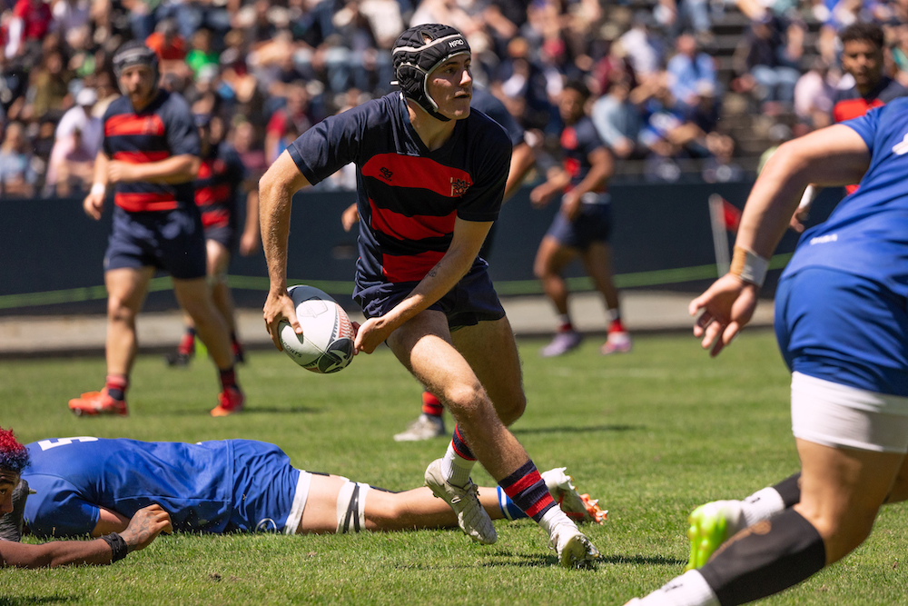 Men's rugby player Hunter Modlin carries the ball while other players lie on the ground, April 2024