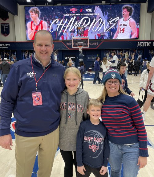 Tom Carroll '04, MA '07 with daughter Aubrey, son Cooper, and wife Stacey Carroll '03 in front of Men's Basketball digital screen