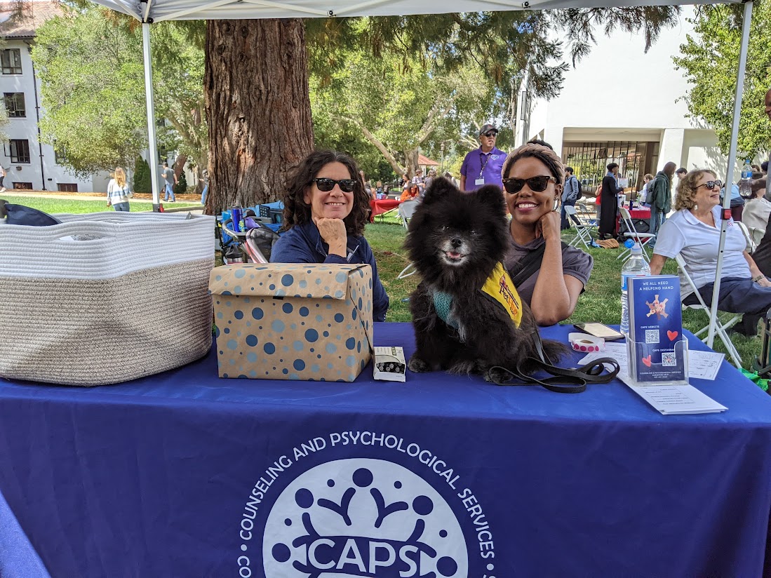 Wellness Fair picture with Joey the therapy dog and two CAPS staff members helping out at the table.