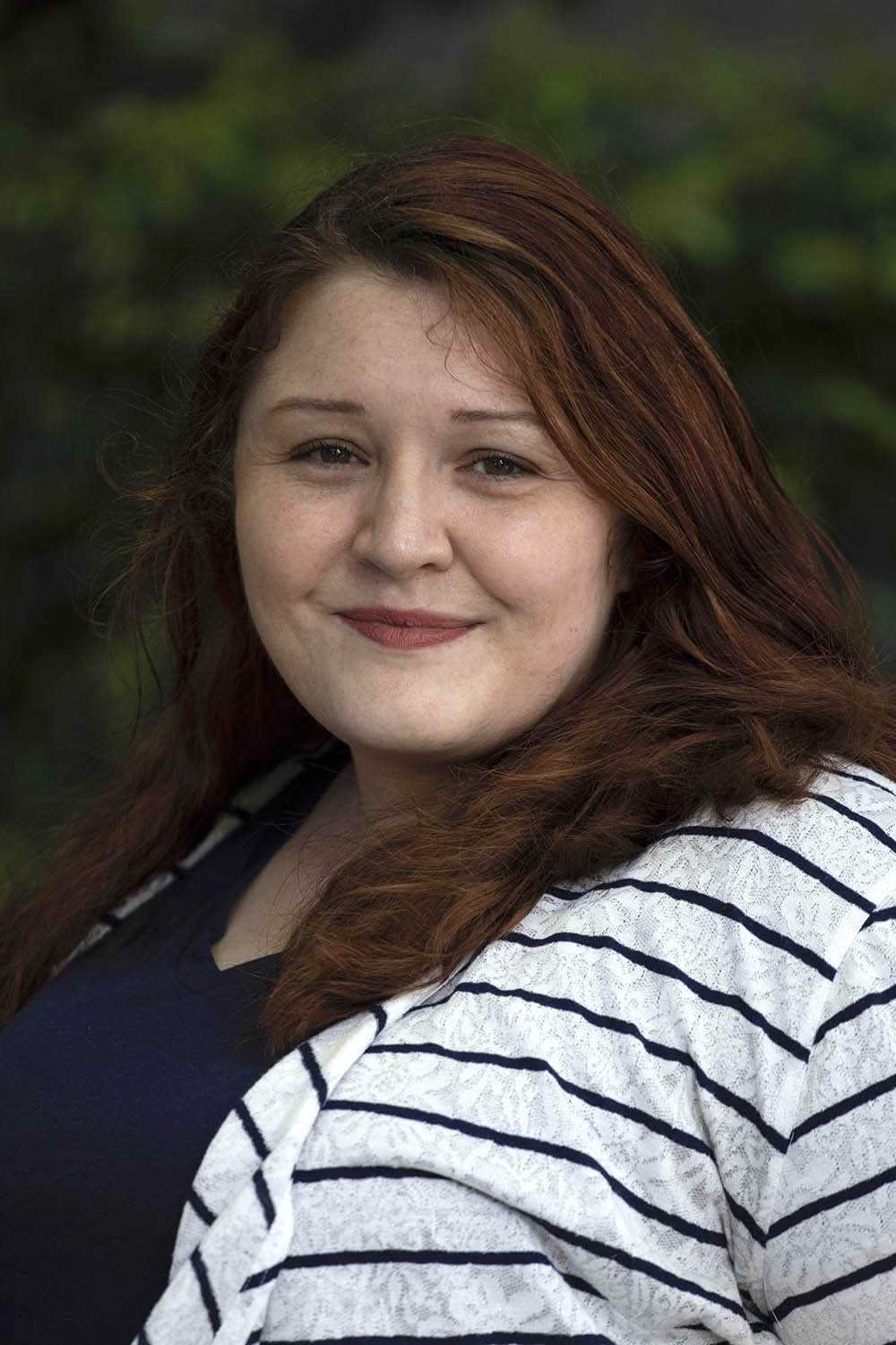 A red-headed woman with a striped black-and-white shirt against faded shrubbery