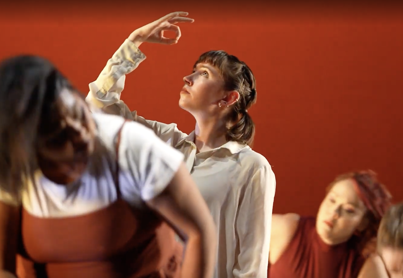 Three dances against a red background the one in the center is wearing a white button down shirt and is looking up at her raised hand