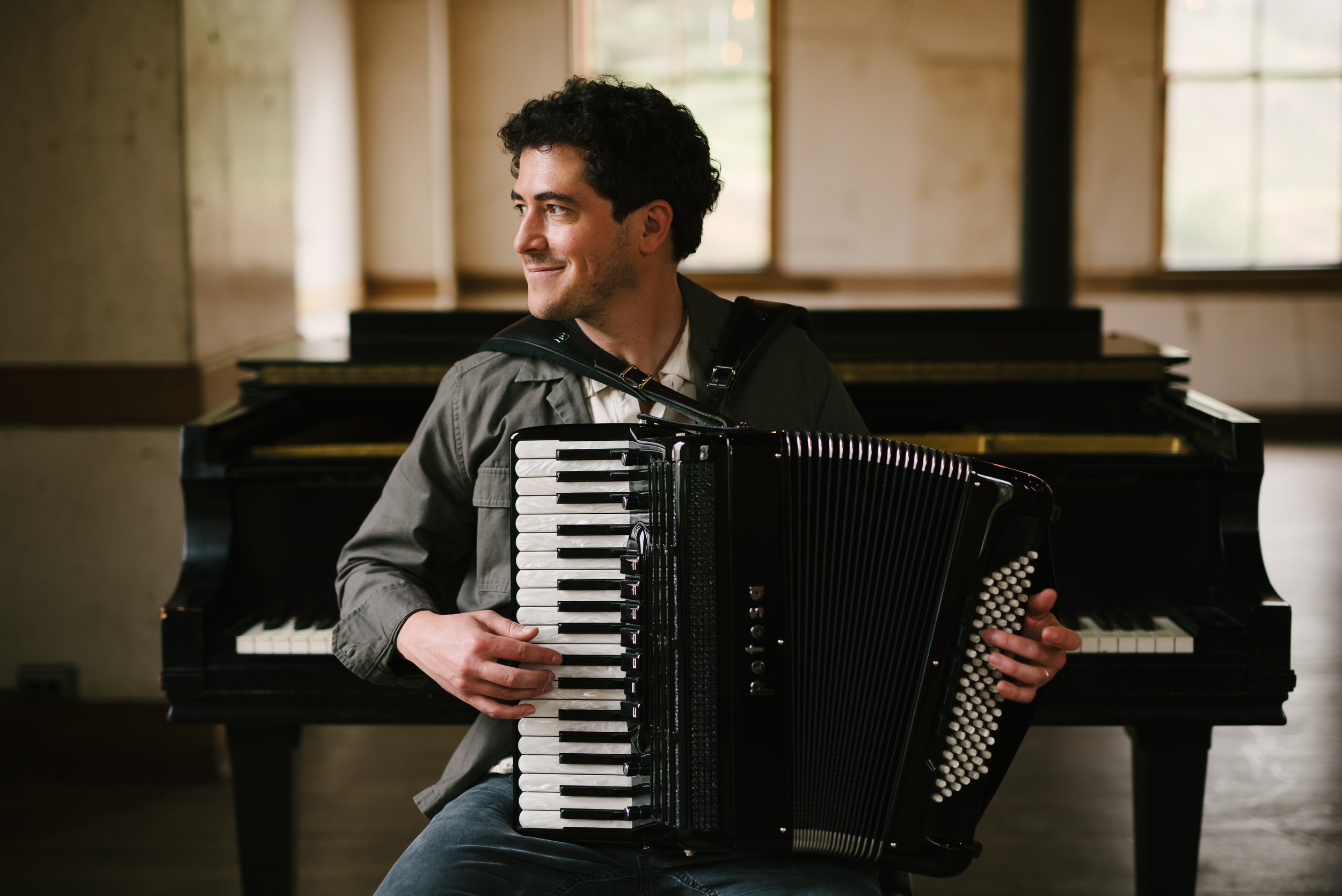 Sam Reider, lookng to the left, holding an accordion. Behind him, a piano.  