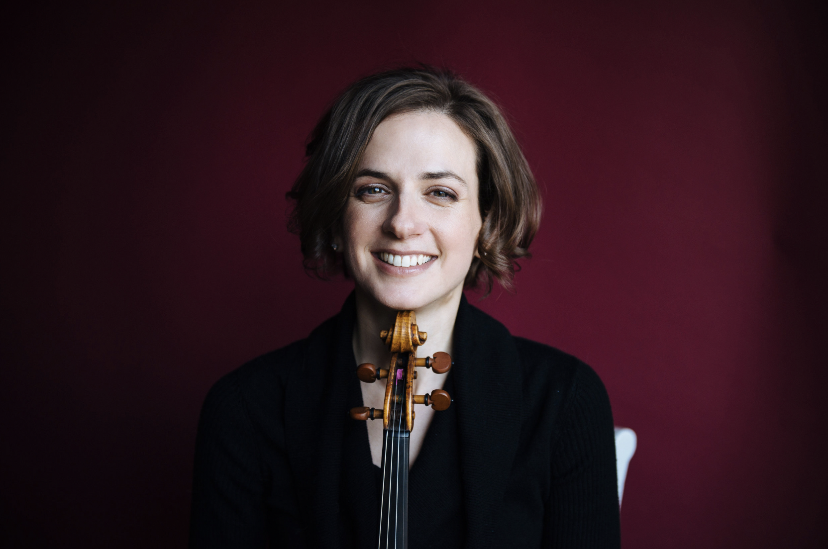 Liana Bérubé against a red background, smiling, and resting her chin on a violin