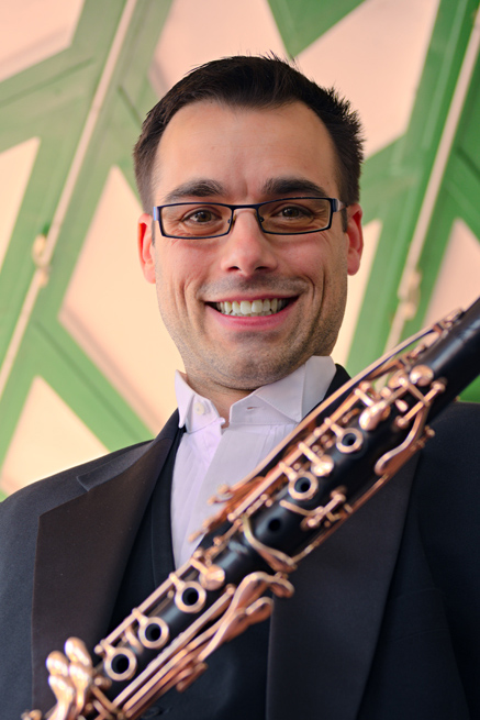 Matthew Boyles against a green and white pattern background, smiling down at camera, whilst nicely dressed and holding his clarinet