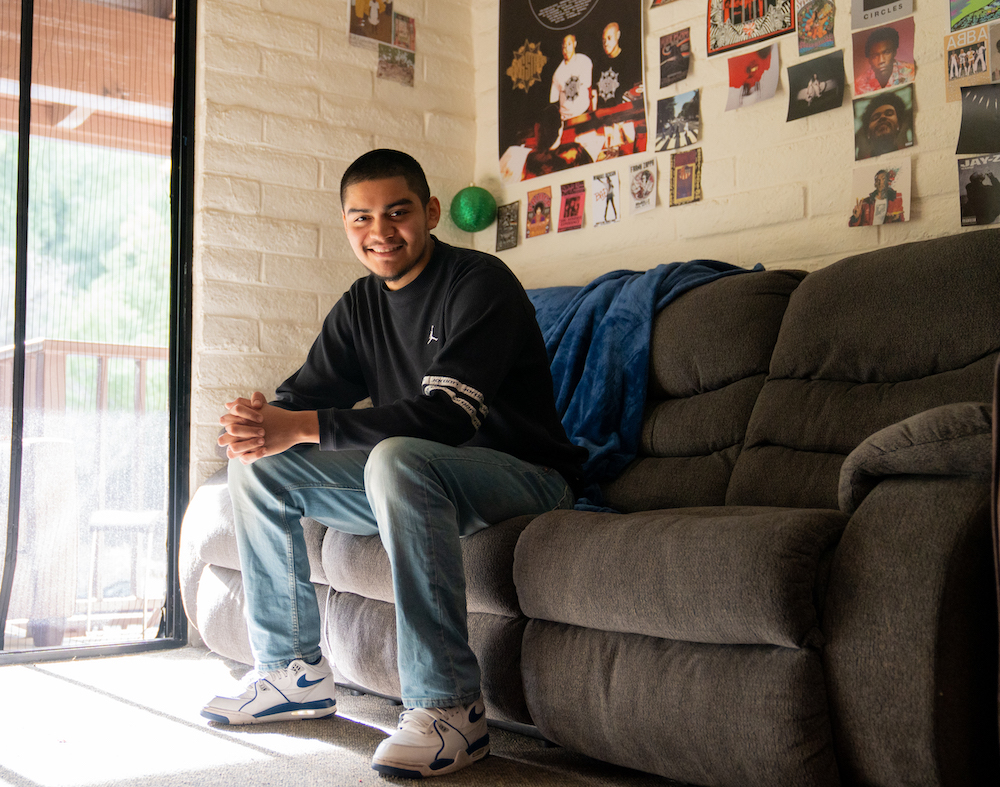 First-generation college student Mat Escalante in his room, seated on a couch with posters in the background.