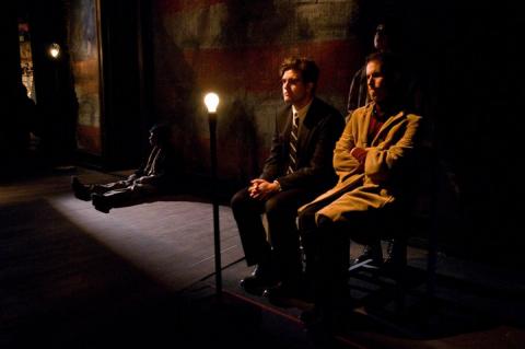 Two people are seated, being lit by a single lightbulb. Their environment (stage) is dreary and dark. In the corner, a man is seated on the floor, hiding in the shadows.