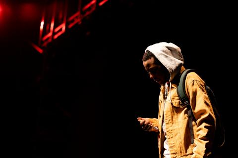 A man in a hoodie looks down while being lit above by a stage light. In his hand, what seems to be a needle. 