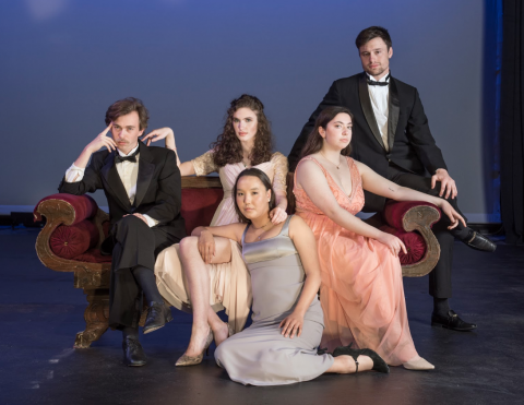 A group photo of fancy-dressed people sitting on a high-end couch on stage. Three women in the middle are wearing dresses while two men at the edge of the couch wear tuxedos. 