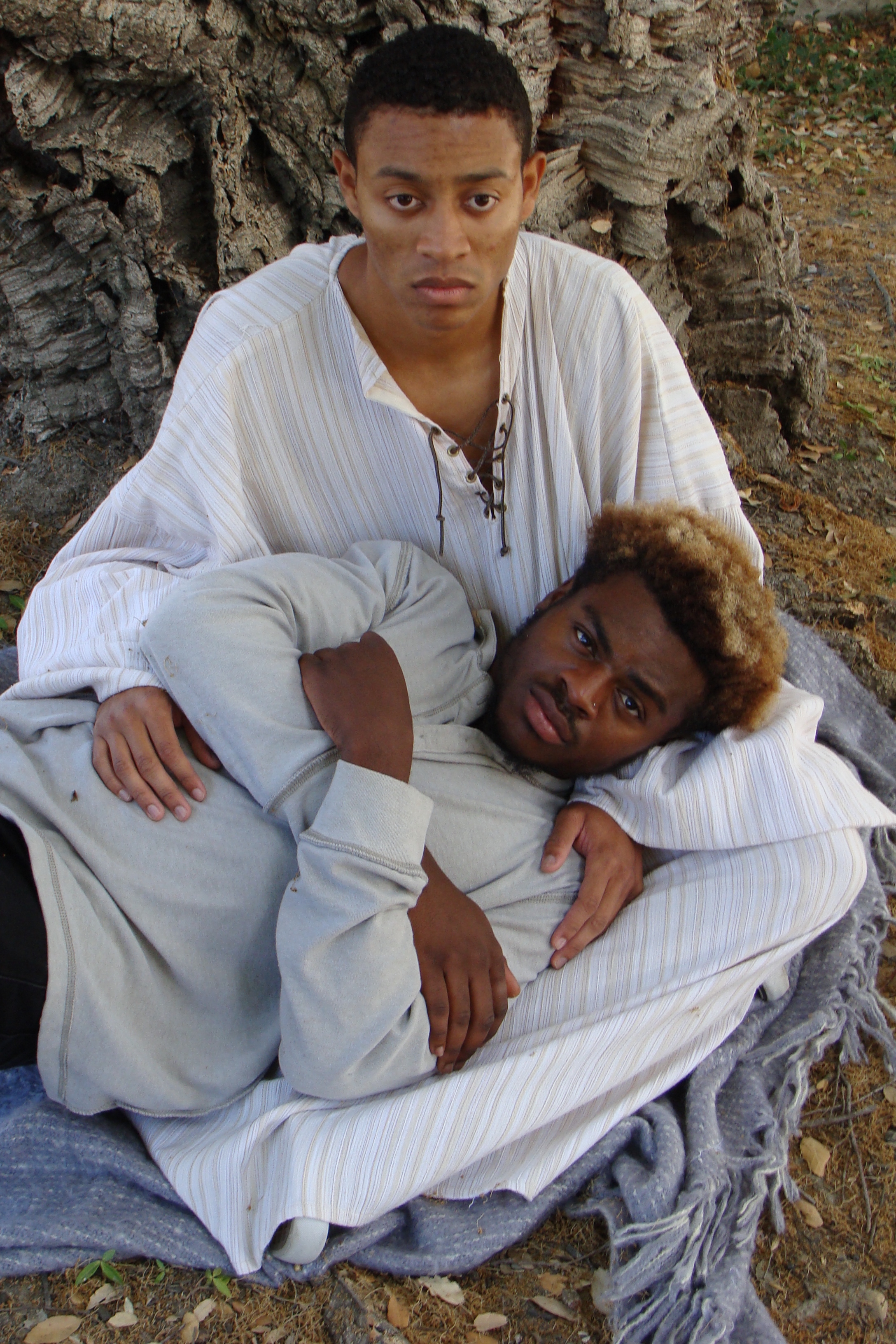 Two Black men wearing white sitting outside, looking up at camera. One of the performers is laying on the other, with his arms crossed. 