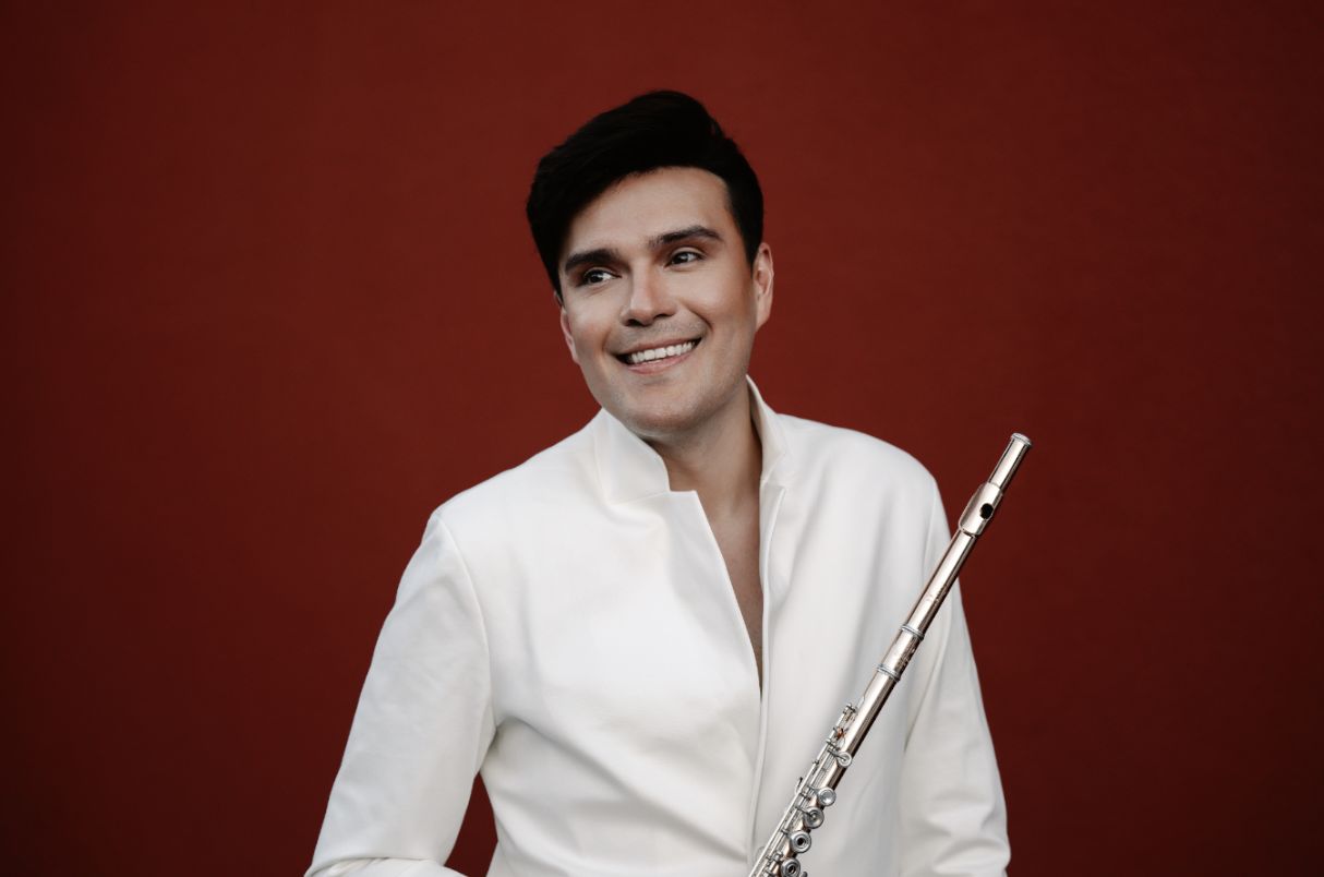 Rayo Furata, holding a flute, smiling and wearing a white dressy shirt with a red background behind him.