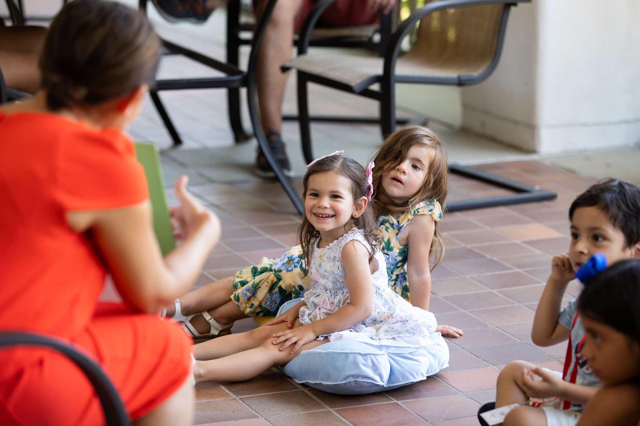 A young child smiels while sitting on the ground while an adult librarian reads a story