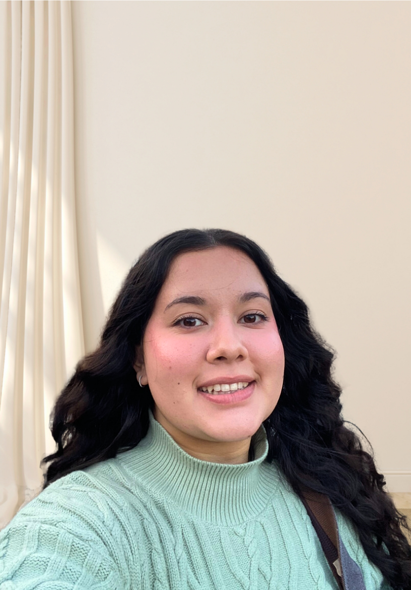 A selfie of Amelia Ortiz, wearing a blue sweater, behind a white wall with a white curtain draped to her left
