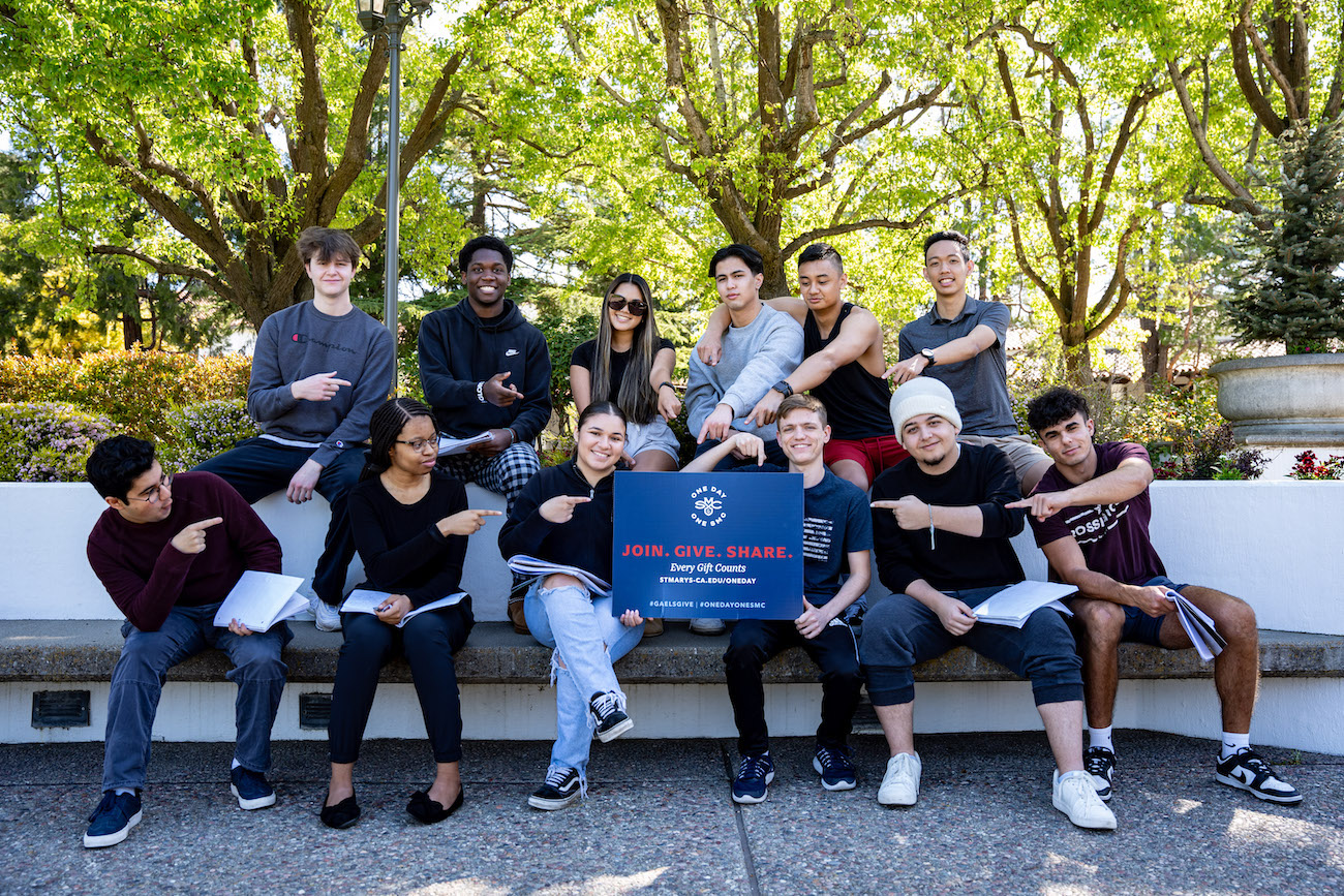 A dozen students point at a One Day One SMC sign on campus