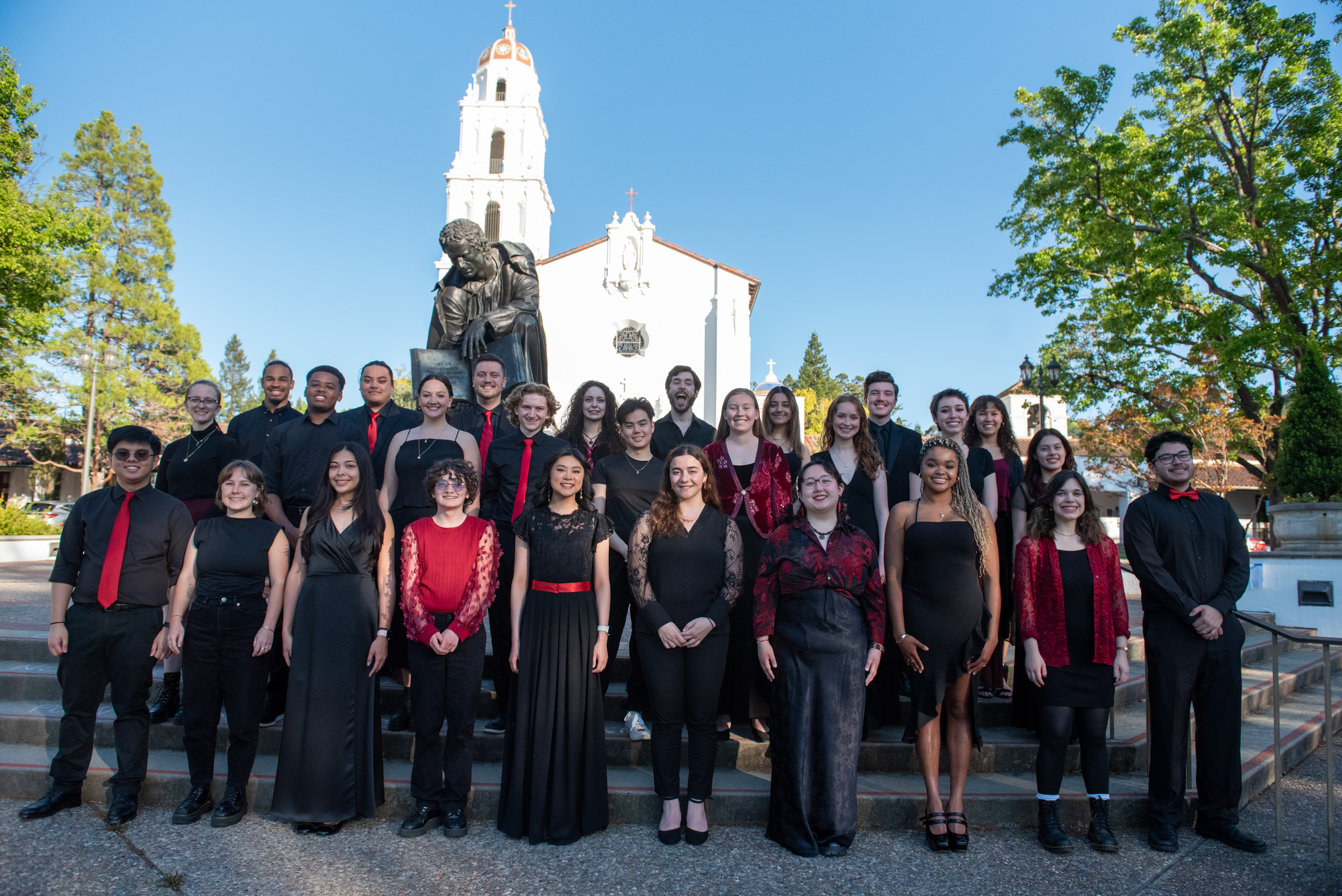 Saint Mary's College choir dressed in black and red stands in front of the SMC Chapel and statue of John Baptist de la Salle with a blue sky above them.