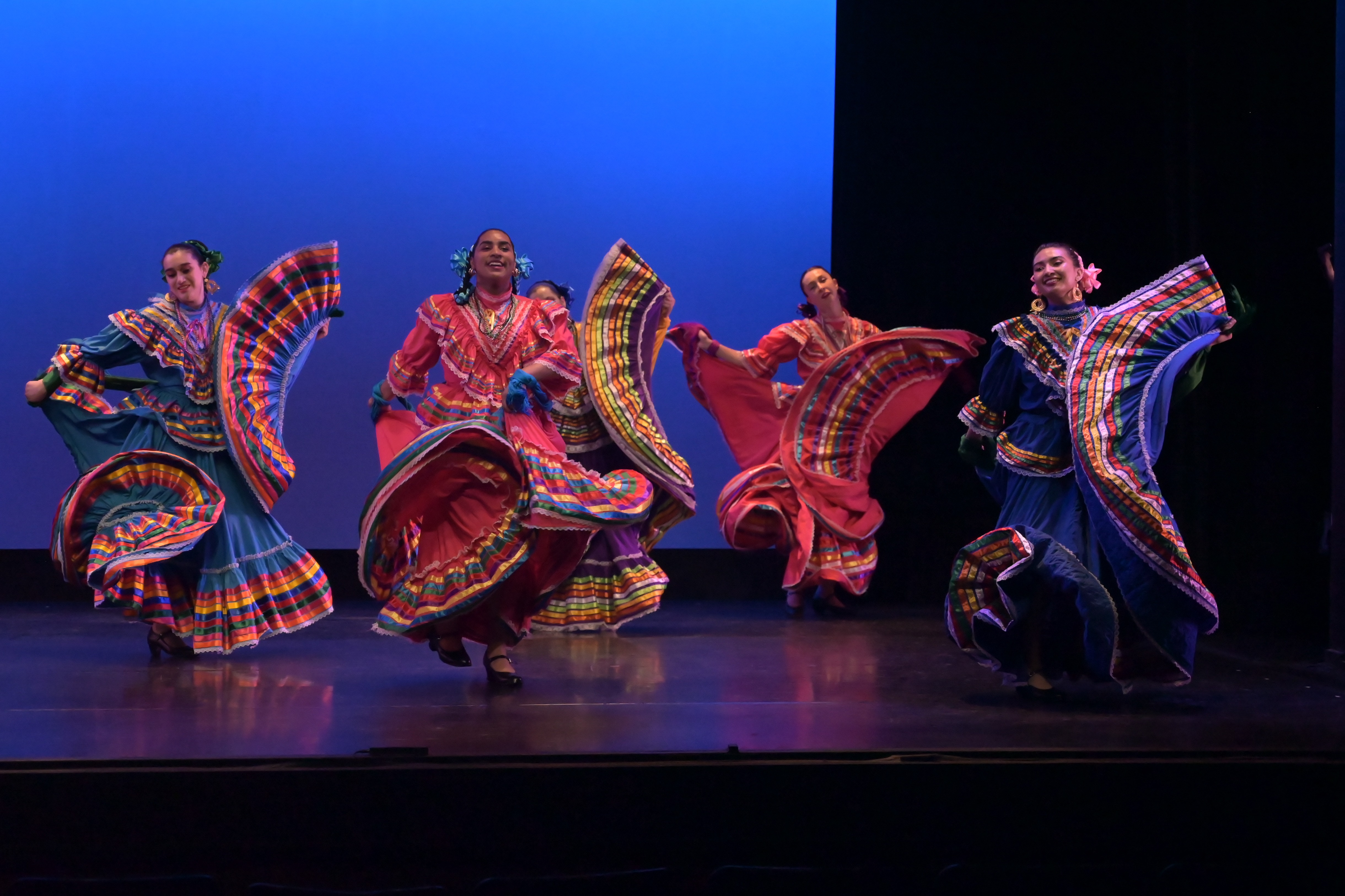 Four BFG dancers in traditional Mexican dance dresses on stage with a blue background