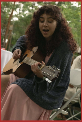 Young woman with long curly auburn hair, Molly Baziuk, plays a guitar and sings while sitting outdoors.