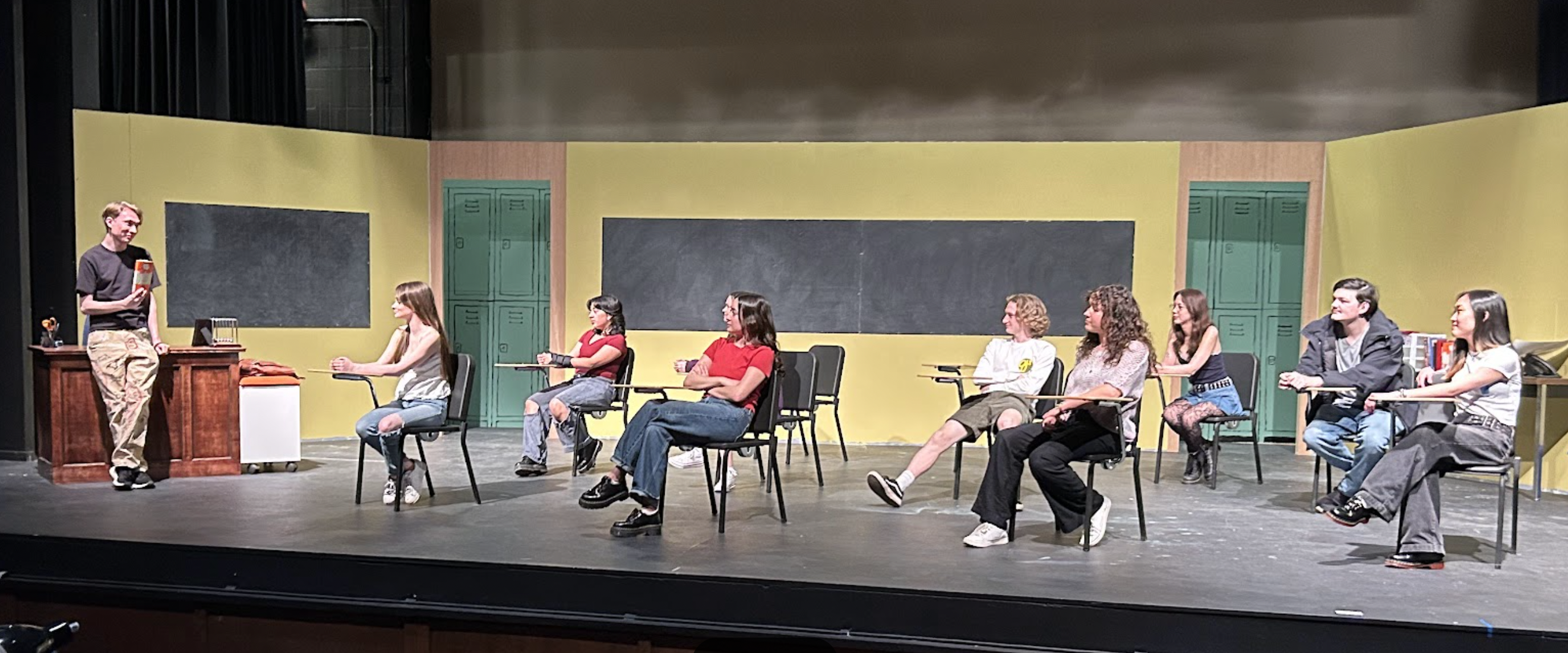 A teacher is holding up a book which the students who are seated at desks in a classroom are looking at. You can tell the class is on a stage as part of a play.