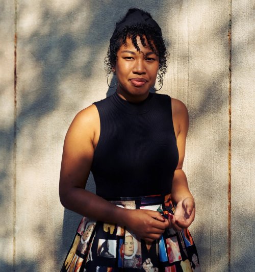 Headshot of woman standing infront of a wooden fence, in a black top and printed skirt