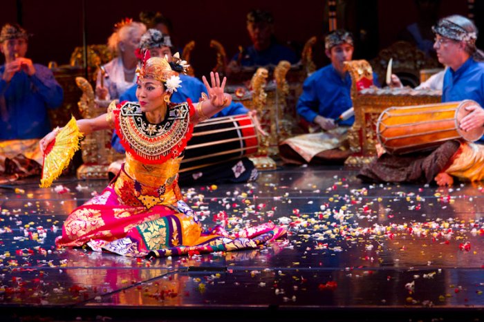 Balinese Dancer in colorful outfit, palms outreached, sitting on the floor with confetti. Instruments being played in the background