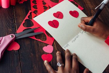Picture of a person making a valentine's day card, scissors used to cut out hearts and a person writing on the card