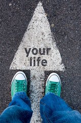 A picture of someones feet, standing on an arrow that it pointing up and says "your life"