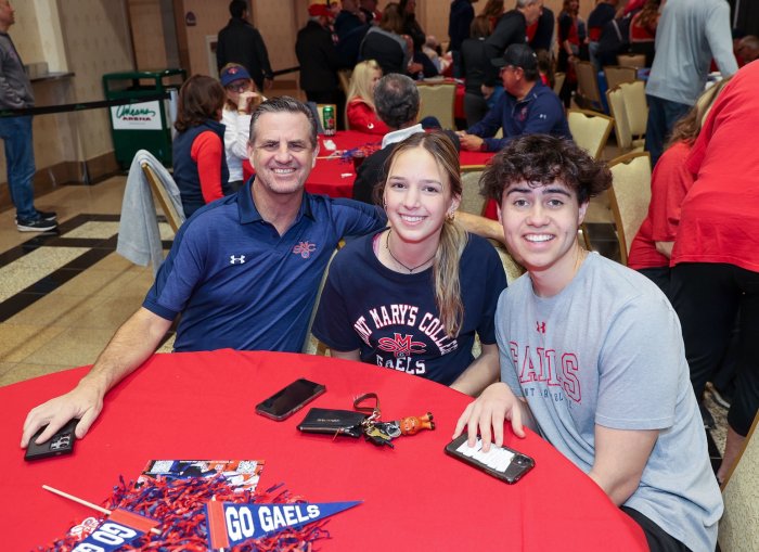 A man and two young adults smile for a photo at a table with many fans behind them