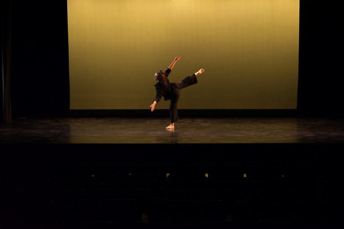 A dancer in black bends forward at the waist and lifts their leg into an attitude position, against a yellow background