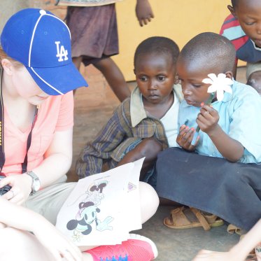 A woman in a blue baseball cap reads to interested young children sitting around her. One holds a flower.