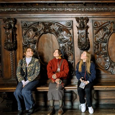 3 students sit on a bench looking up against an intricately carved wood wall.