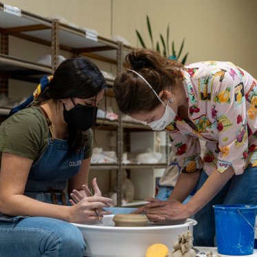 A student and professor lean over a pottery wheel in a ceramics studio , forming the clay.