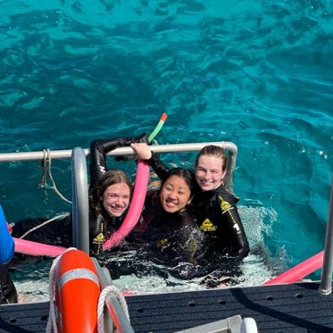 Three students in scuba gear pose for a photo hanging off a boat in bright turquoise water.