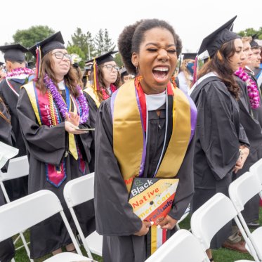 A graduating student yelling while holding a hat that says Black Native