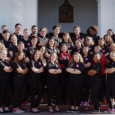 A group photo of the 2019 choir group in front of the chapel, wearing black shirts and pants, their arms crossed.