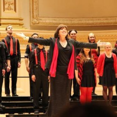 Dr Julie Ford, dressed in black-and-red, extends her arms to the crowd while the choir stands behind her, arms to their sides