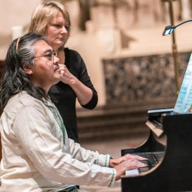 Lino Rivera and Renée Witon at the piano in the chapel. Lino is playing the piano and Renée stands next to him, reading the sheet music alongside him