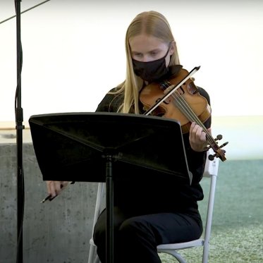 A violinist outside wearing a black mask during the COVID-19 pandemic. She is reading music from her music stand