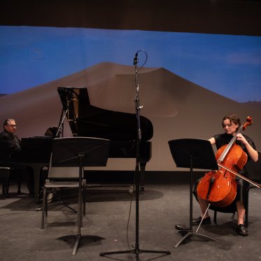 Two musicians, one piano player and one cellist, performing on stage in LeFevre theatre against an animated background of a desert landscape at twilight. There is a large sand dune in the middle of the screen. 