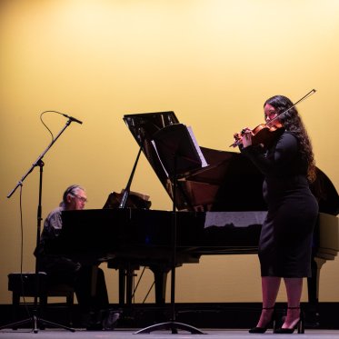 A string instrumentalist performs on stage at LeFevre Theatre while Lino accompanies them on a piano
