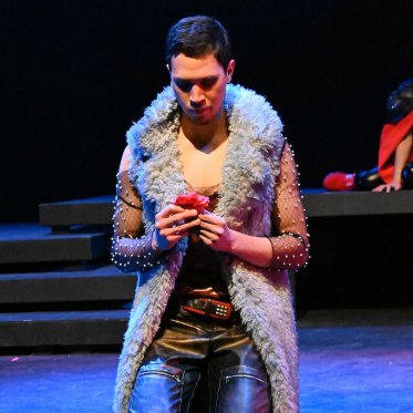 A photo of a man wearing a gray scarf, bedazzled sleeves, on their knees and holding a pink rose -- a still from "A Midsummer Night's Dream"