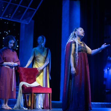 Send It To The Moon -- a woman in ancient Greek robes, black and maroon, extends her hand out into the audience. Two women in the darkness, one dressed in red and the other yellow, stand near a red chair. 