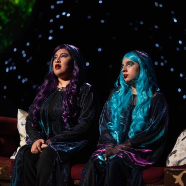 Send It To The Moon -- two women in purple and blue hair look out past the stage, with a light shining on them. Behind them, a cosmic backdrop. 