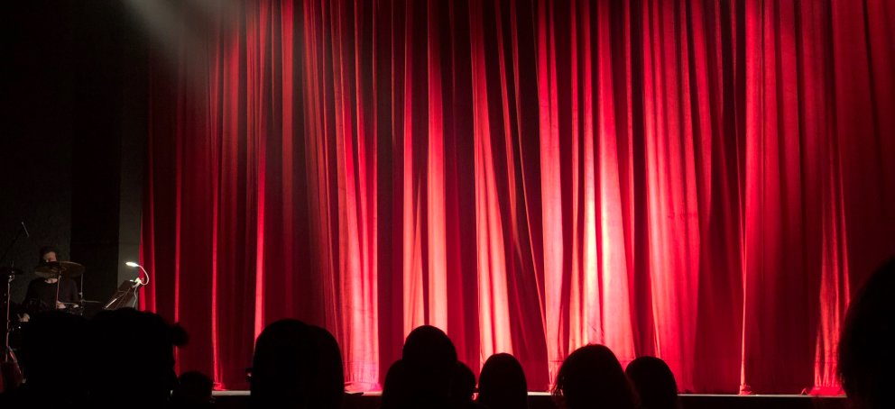 Red curtain lit up by stage lights with shadows of audience in the foreground.