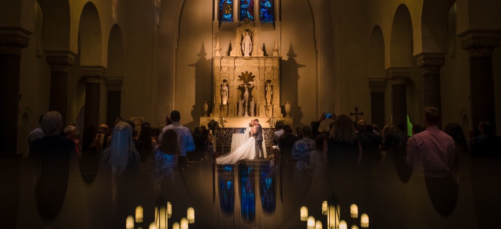 A wedding couple celebrate their first kiss in the SMC Chapel surrounded by their guests. The lights are dim and the reflection of the chandeliers in the tile floor appears like candlelight.