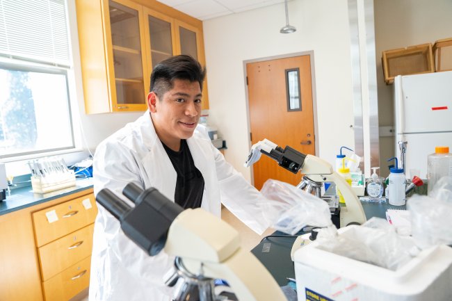 male student standing in a lab coat over microscopes within a lab classroom surrounded by lab equipment, smiling at the camera