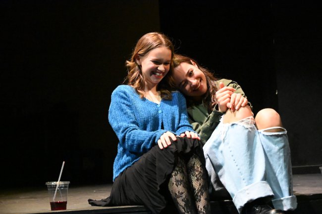 Two female friends sit side by side in a scene on stage