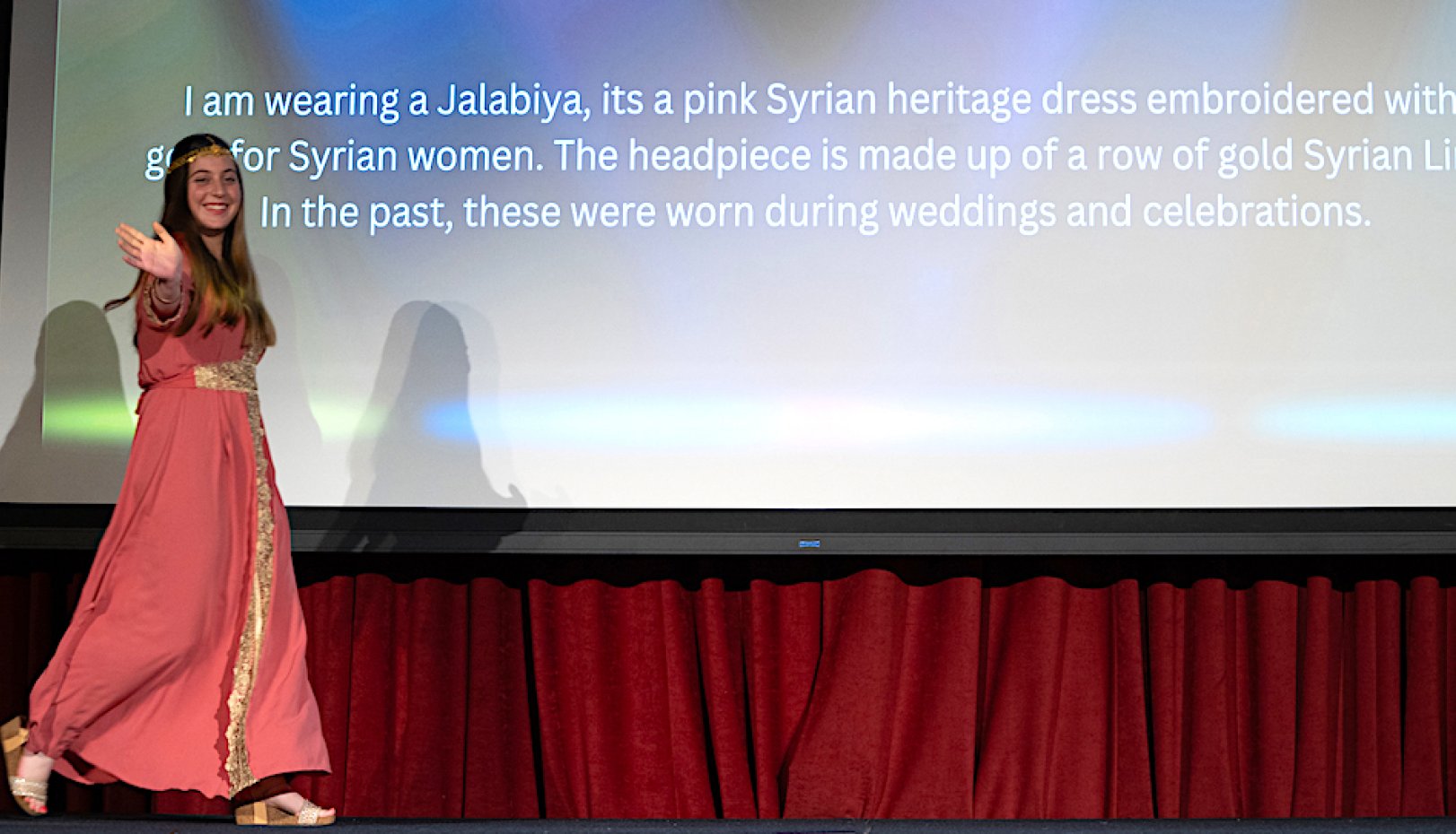 Vanessa Al Nader wears a traditional Syrian dress and walks in front of text explaining it