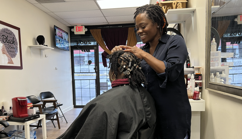 A woman sits in a salon chair while a hair stylist works on her hair.