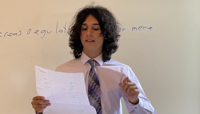 Young person in white shirt and purple tie holds a white paper with writing as he stands and speaks from those notes.
