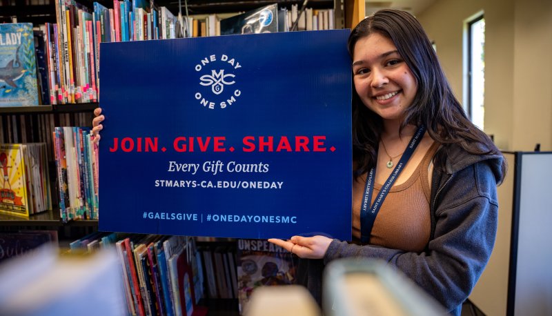 a smiling student with long brown hair holding a One Day One SMC sign